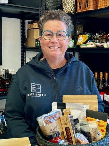 A smiling woman with short curly gray hair and glasses, wearing a navy blue zip-up hoodie with 'GIFT SMITH' printed on the chest, holds a large woven gift basket filled with packaged snacks and treats in a retail shop setting.